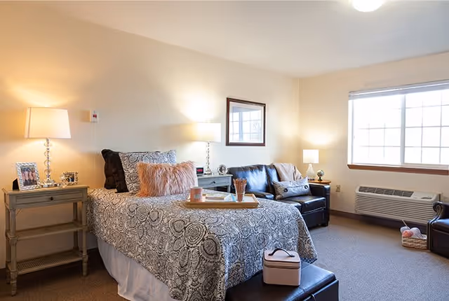 A cozy bedroom in an assisted living facility featuring a single bed with patterned bedding and decorative pillows. Beside the bed are two nightstands with lamps, a framed photo, and small decor items. A black leather loveseat and a small table lamp are positioned near a large window with blinds. The room has neutral-colored walls and carpeted flooring, with an air conditioning unit below the window.