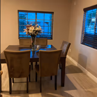 A dining area with a dark wooden table and four brown upholstered chairs. A vase with a bouquet of flowers is placed in the center of the table. Two windows with wooden blinds are visible on the walls, letting in some natural light.