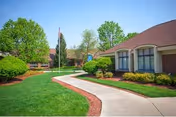 Curved concrete walkway through a manicured lawn leading to a single-story brick rehabilitation center with shrubs and an American flag.