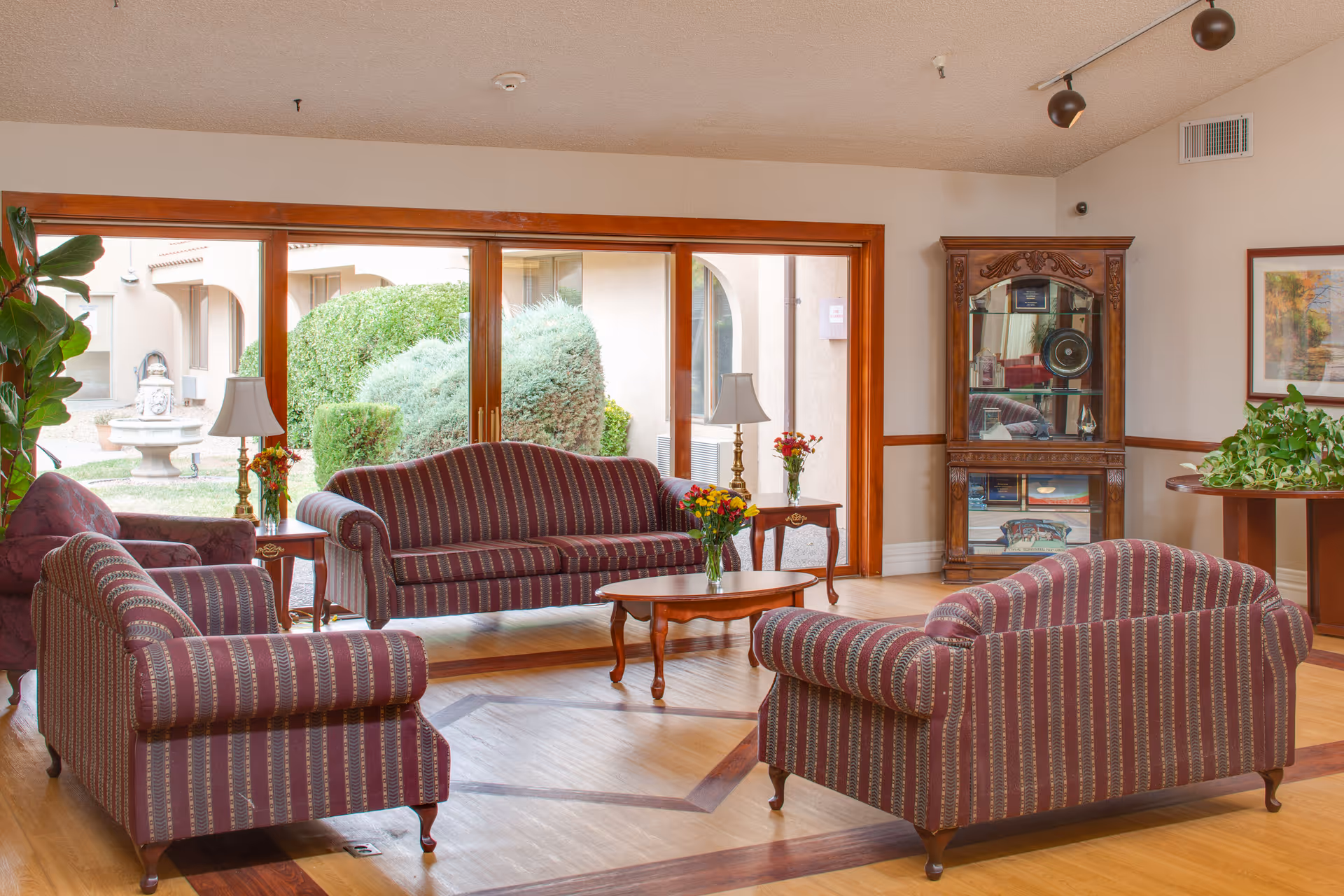 Bright lounge with striped sofas and armchairs arranged around a coffee table in front of large windows overlooking landscaped shrubs.