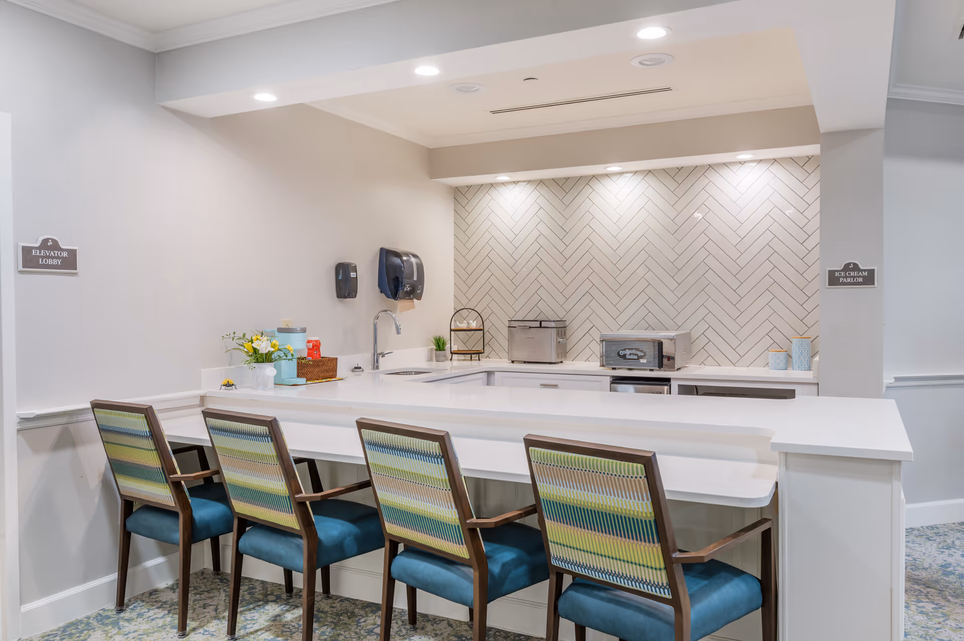 A clean and modern ice cream parlor counter area with four chairs featuring blue seats and patterned backs. The counter is white with a sink, a small flower arrangement, and some appliances against a wall with white herringbone tile. Signs on the walls indicate directions to the Elevator Lobby and Ice Cream Parlor.