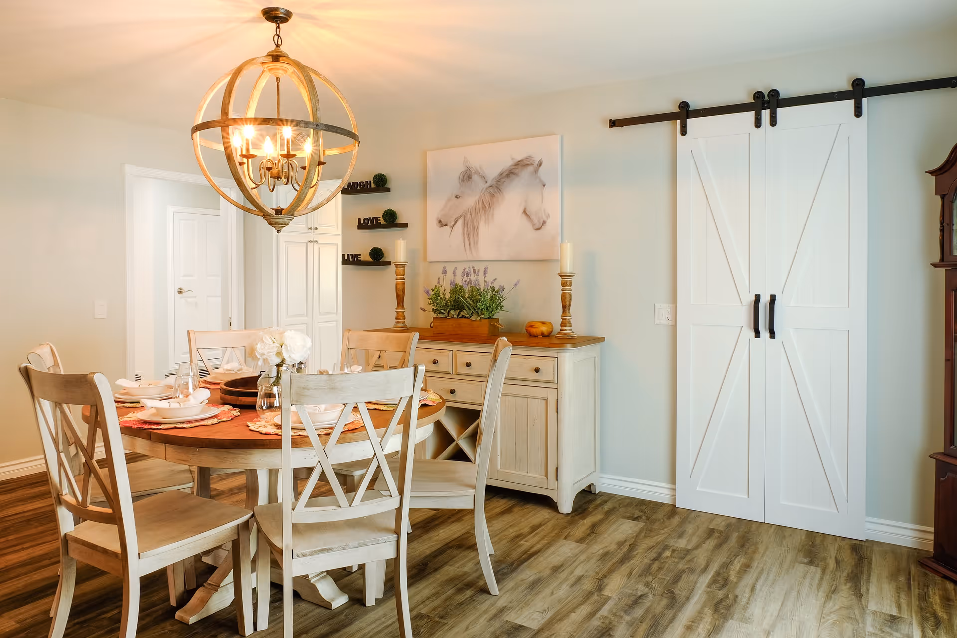 A cozy dining room with a round wooden table set for four, surrounded by four wooden chairs with cross-back design. Above the table hangs a spherical chandelier with multiple light bulbs. Against the wall is a sideboard with decorative items including candles, a plant, and a painting of two white horses. To the right, there are white sliding barn doors on a black metal track. The room has wood flooring and light-colored walls.