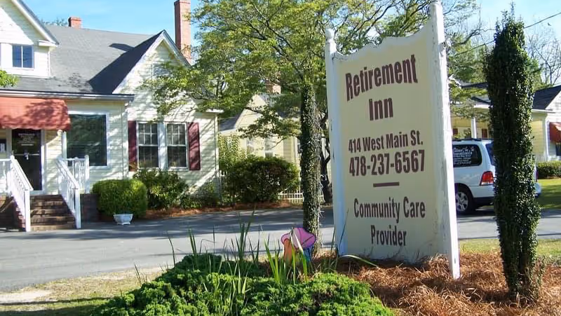 Exterior view of a white building with a red awning and a sign in front that reads 'Retirement Inn, 414 West Main St., 478-237-6567, Community Care Provider'. There are trees and shrubs around the sign and building.