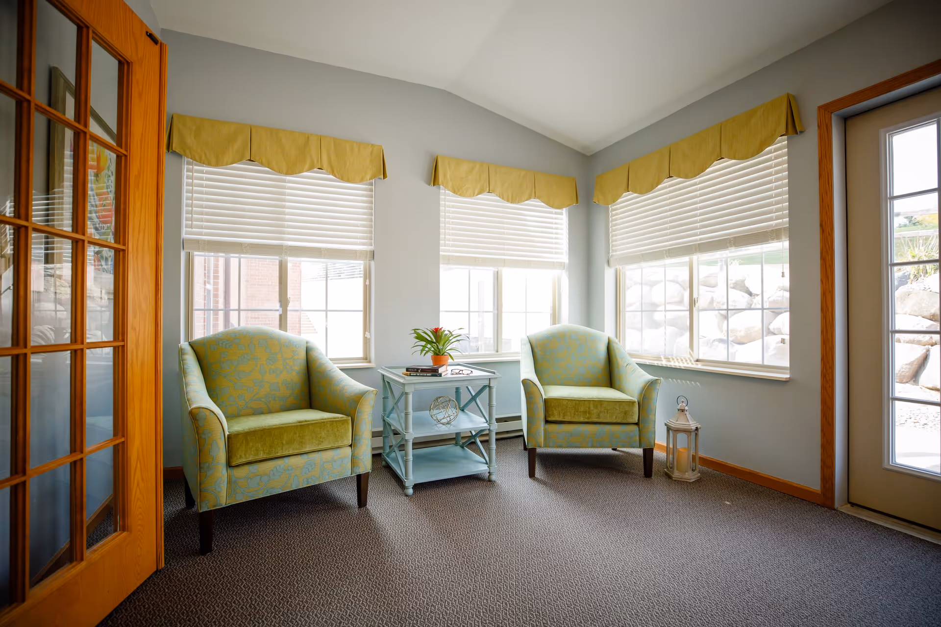 Sunlit seating area with two green patterned armchairs flanking a small blue side table in front of three windows.