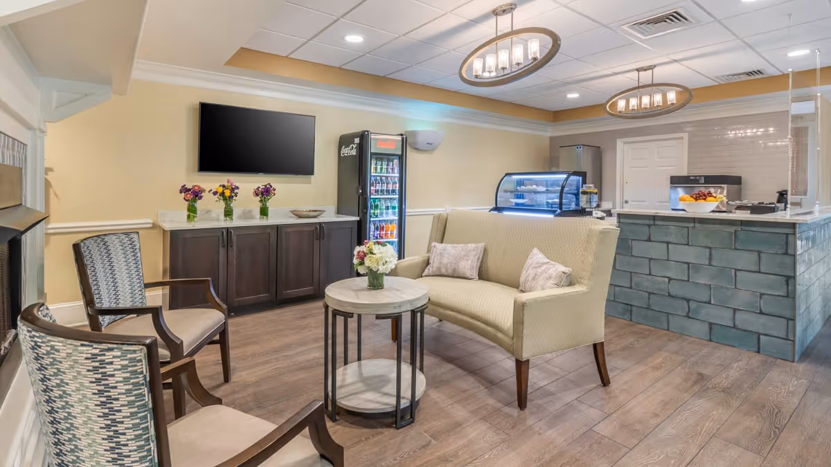 A well-lit common lounge with a sofa, chairs, a wall-mounted TV, a beverage fridge, and a service counter with display case.