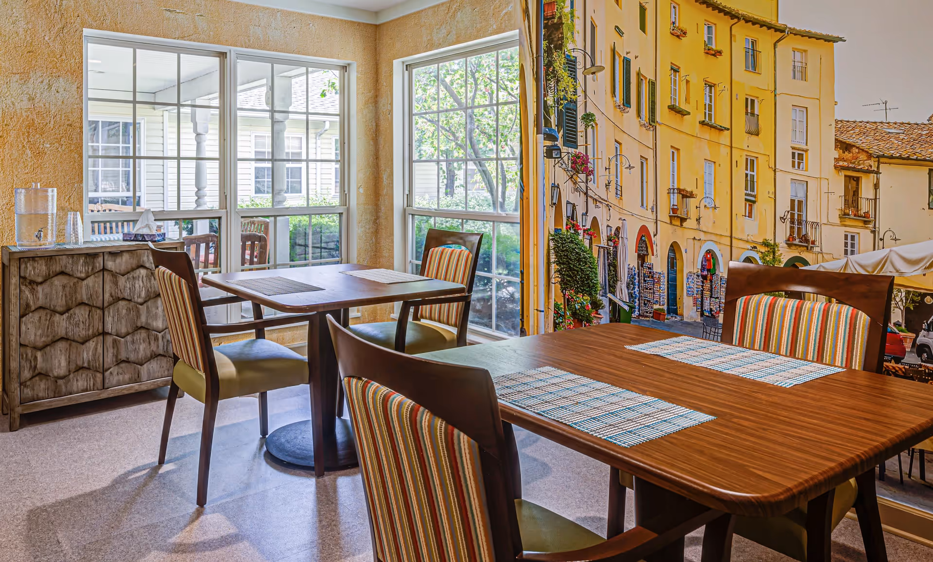A cozy dining area with two wooden tables and four chairs featuring striped upholstery. The room has large windows letting in natural light and a sideboard with a water dispenser and cups. One wall is decorated with a large mural of a European street scene with yellow buildings and outdoor market stalls.