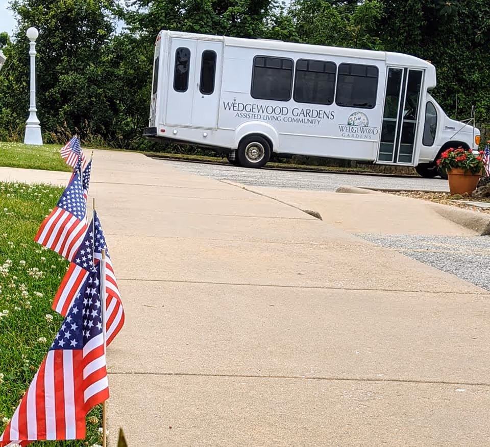 A white shuttle bus with the text 'Wedgewood Gardens Assisted Living Community' parked on a driveway. In the foreground, several small American flags are planted in the grass along the sidewalk. There is greenery and a white lamp post in the background.
