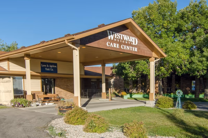 Exterior view of Westward Heights Care Center showing the entrance with a covered porch, a bench, and surrounding greenery including trees and shrubs under a clear blue sky.