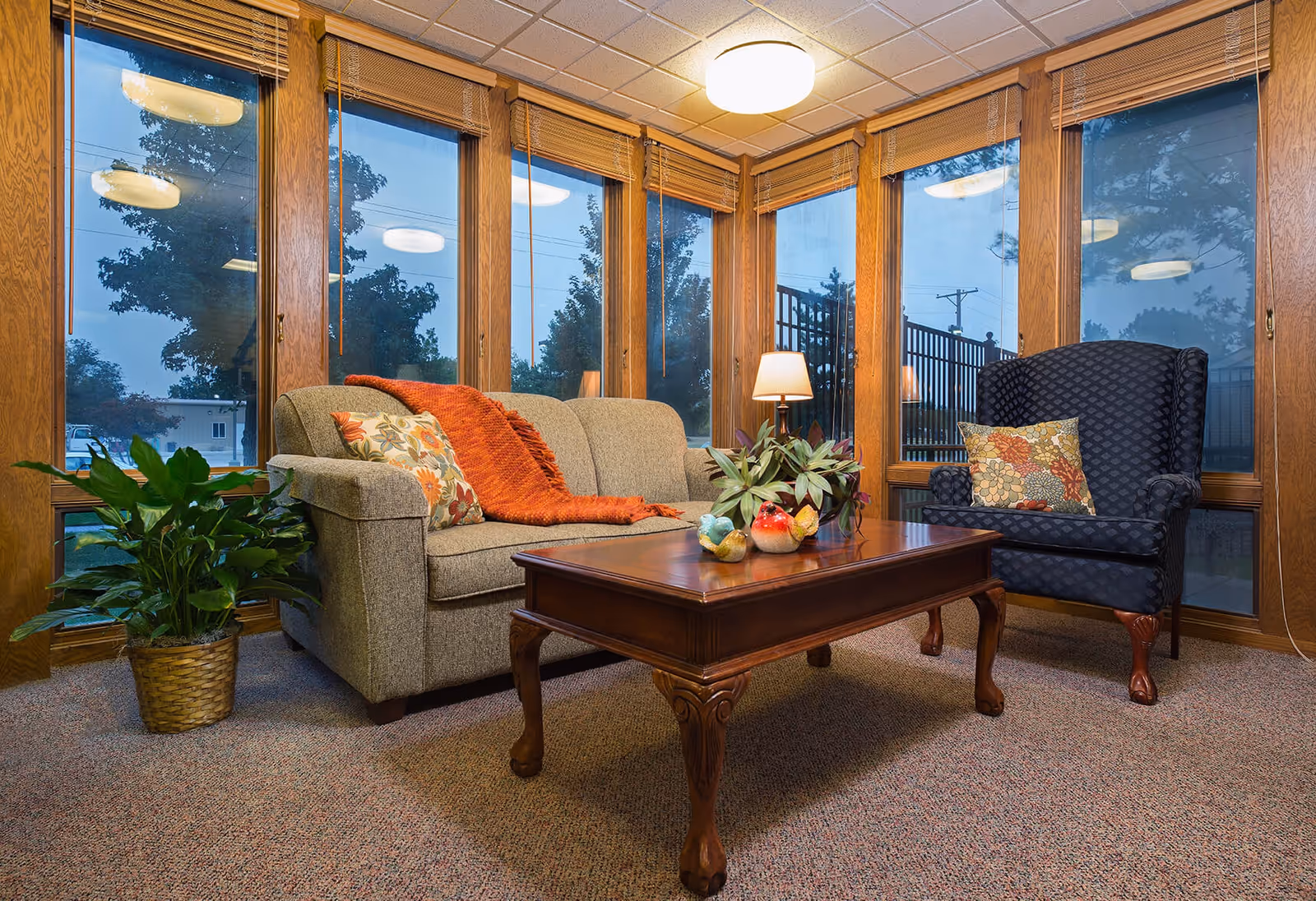 A cozy sitting area with a beige sofa adorned with floral and orange throw pillows and a matching orange throw blanket. Next to the sofa is a dark blue armchair with a floral pillow. A wooden coffee table with decorative items and a potted plant is in front of the seating. The room has large windows with wooden frames and blinds, allowing a view of trees and an outdoor fence. The carpet is a neutral tone, and there is a ceiling light and a table lamp providing warm lighting.