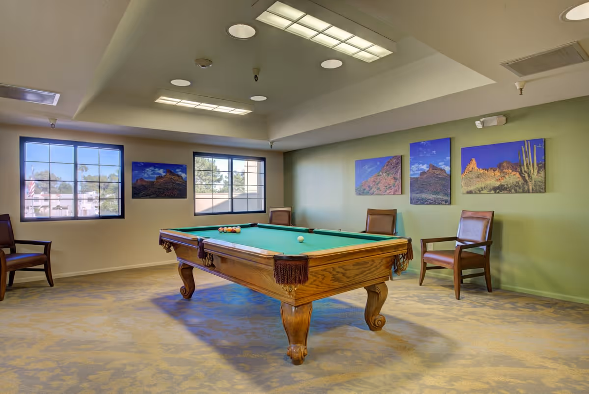 A well-lit room with a pool table in the center, surrounded by four wooden chairs with leather seats. The walls are painted light green and beige, decorated with three landscape paintings of desert scenes. Two large windows allow natural light to enter the room.