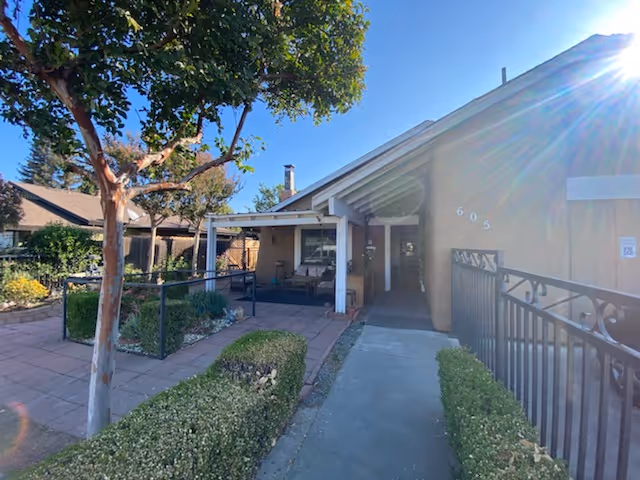Front entrance of a single-story care home with a covered porch, walkway, hedges and a tree under a sunny sky.