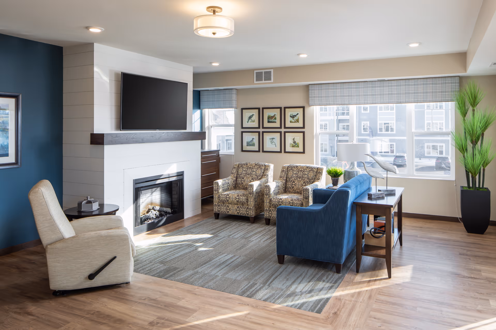 Bright communal living room with a fireplace, wall-mounted TV, sofa, and several armchairs.