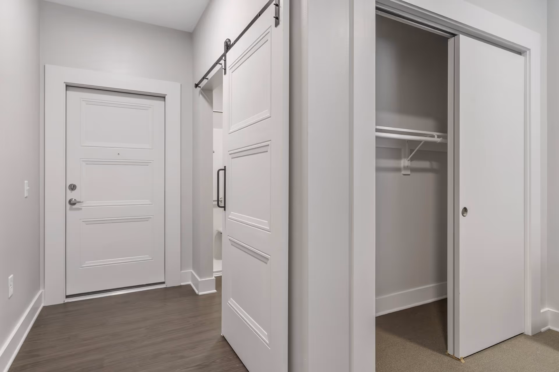Interior view of a hallway in a senior living facility showing a white entrance door, a sliding barn door partially open revealing a bathroom, and a closet with sliding doors and a hanging rod inside. The walls are light gray and the floor is a dark wood laminate.