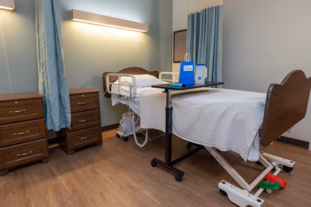 A hospital-style patient bed in a furnished room with a dresser, blue curtains, and medical equipment.
