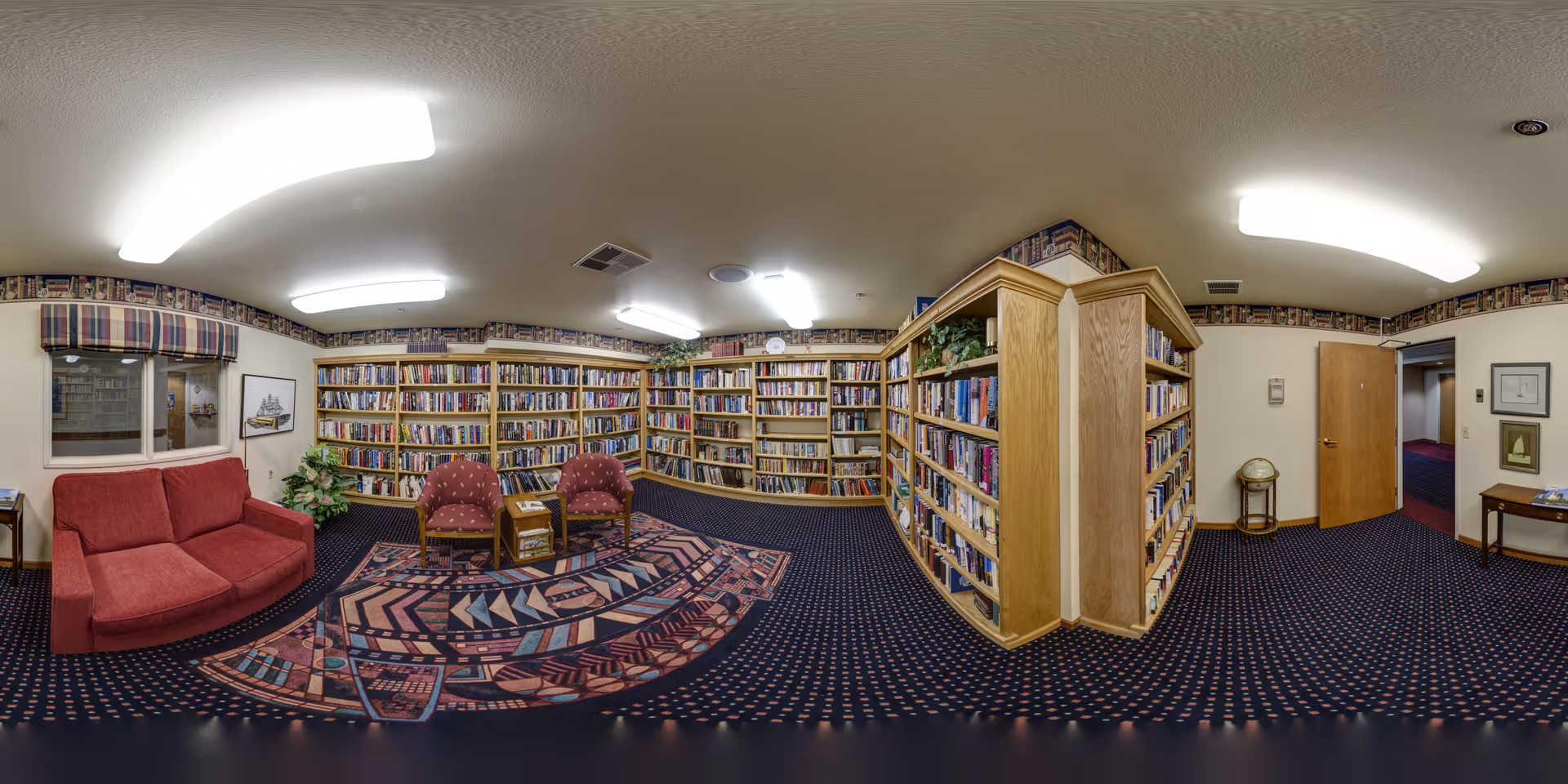 Carpeted communal library room with floor-to-ceiling bookshelves, upholstered seating, and a patterned rug.