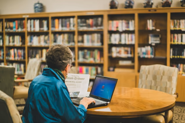 An elderly person wearing a teal jacket is sitting at a round wooden table in a library or common area, using a laptop. Bookshelves filled with books are visible in the background.