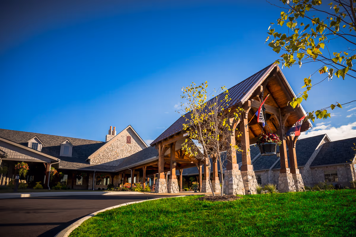 Exterior view of The Inn at Walnut Trail, showing a stone building with a covered entrance supported by wooden beams, surrounded by green grass and small trees under a clear blue sky.