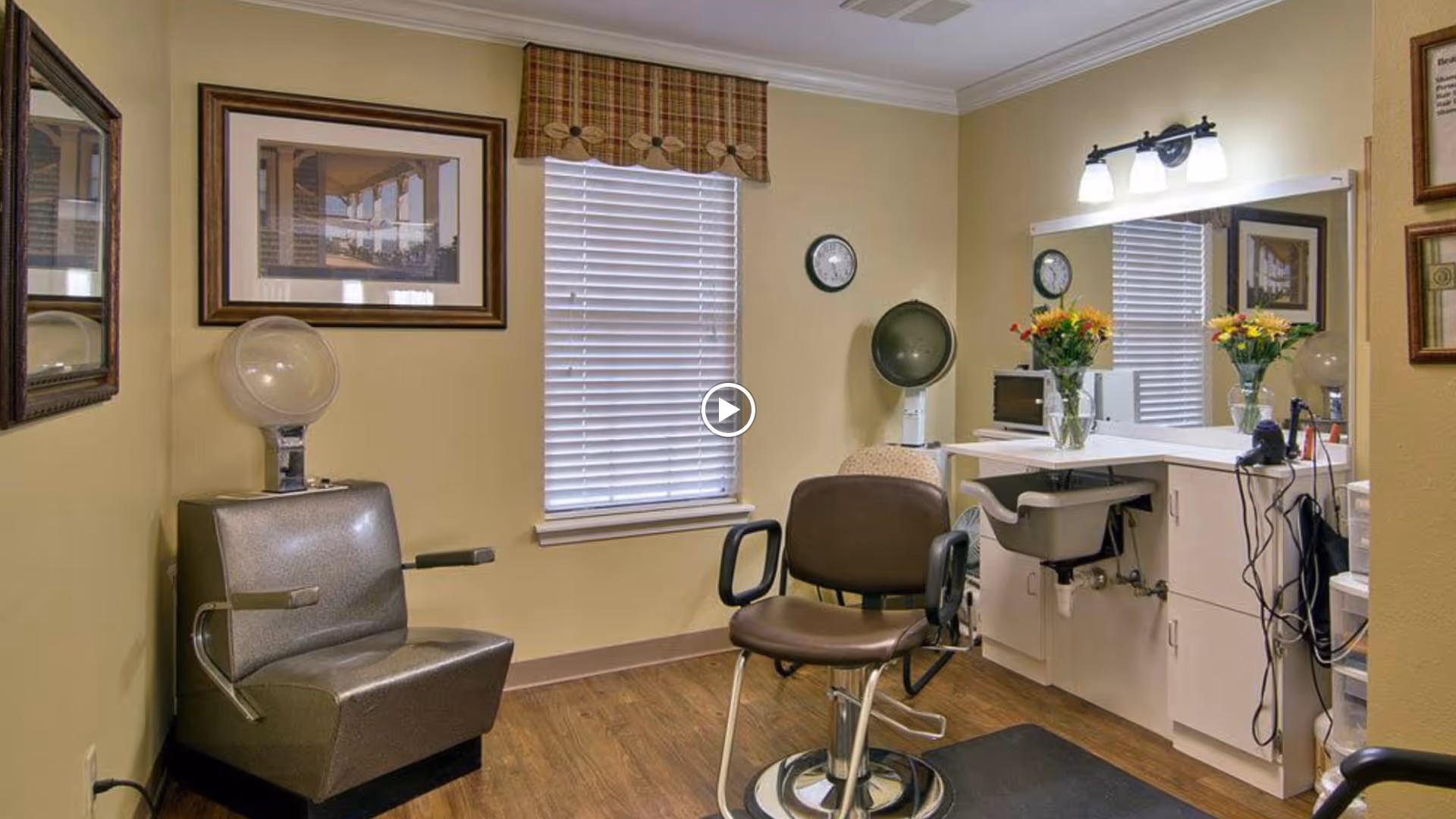 Interior of a salon or beauty room with two vintage hair dryer chairs, a large mirror above a white counter with a sink, flowers in vases, and framed pictures on the beige walls.