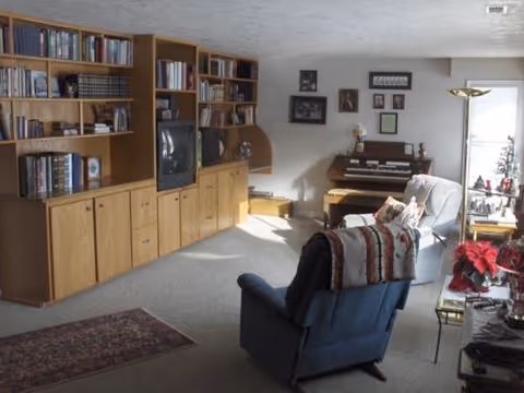 A cozy living room with wooden bookshelves and a TV on the left, two recliner chairs in the center, and a small organ and lamp against the far wall.