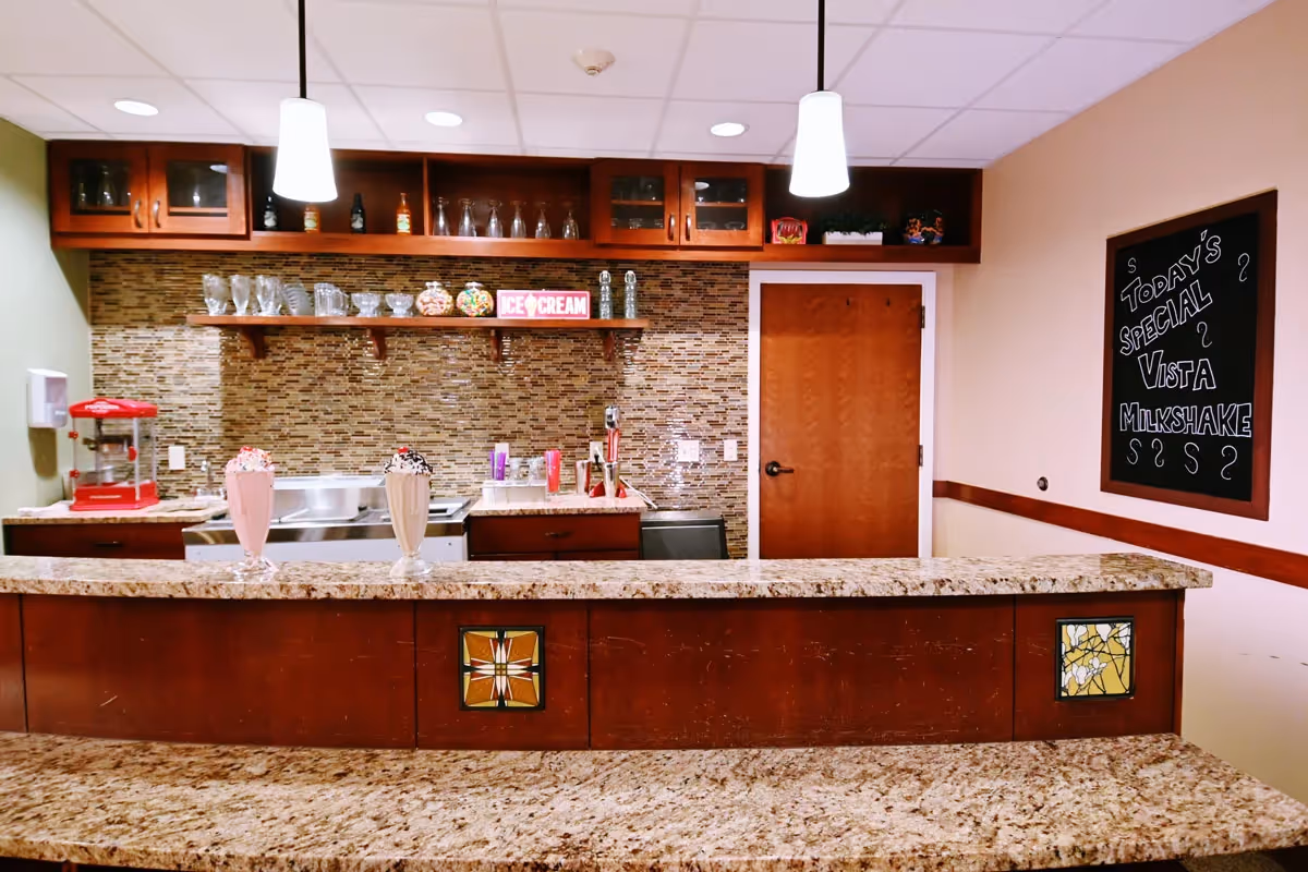A cozy indoor snack bar/kitchenette with a granite countertop, pendant lights, glassware on shelves, and a chalkboard announcing today's special milkshake.