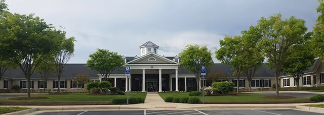 Front exterior view of Spring Arbor of Winchester facility with a wide driveway, landscaped lawn, trees, and a building entrance featuring columns and a cupola on the roof under a cloudy sky.