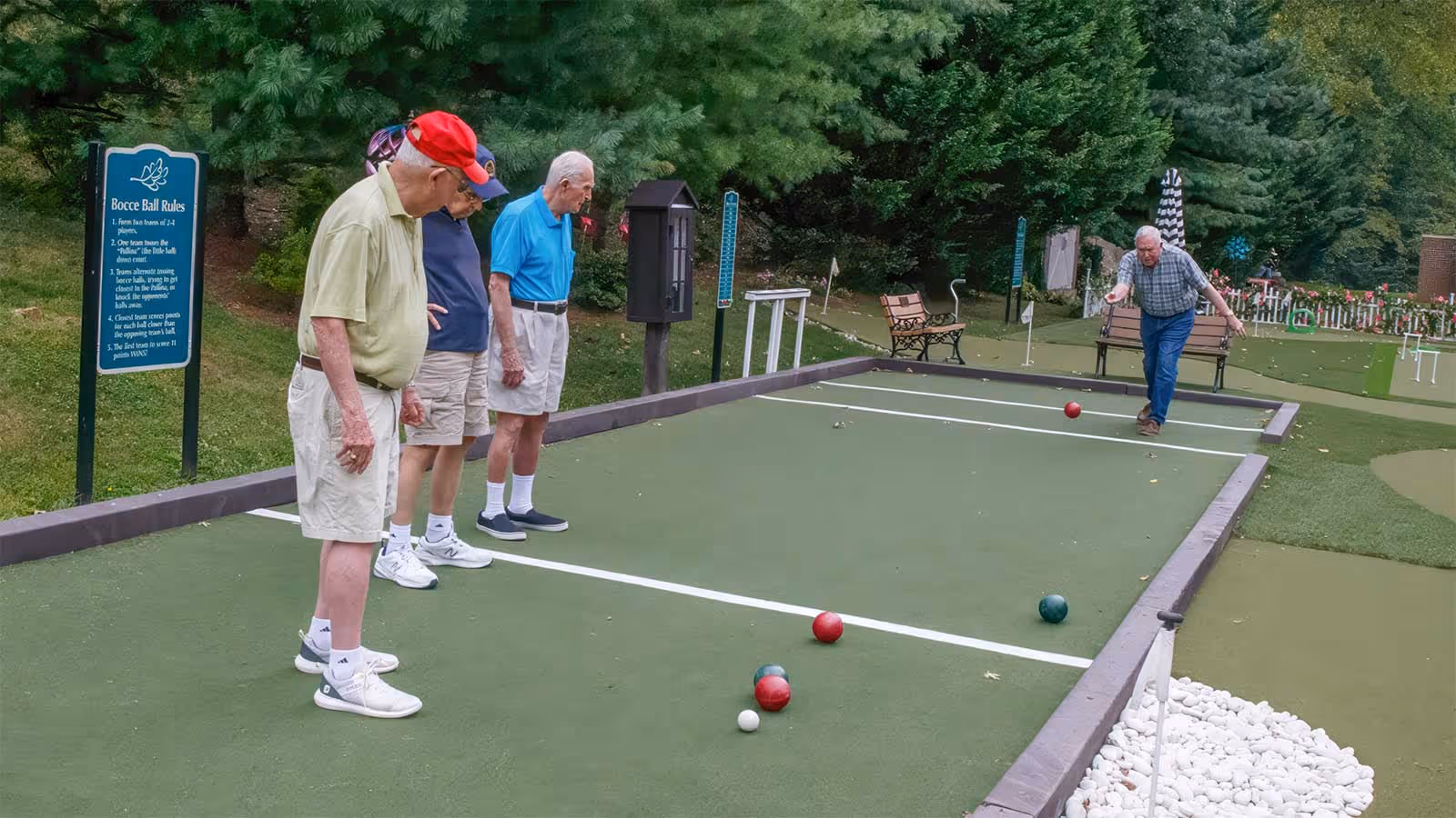 Four elderly men playing bocce ball on a bocce court surrounded by greenery. One man is in the process of throwing a bocce ball while the other three watch. There is a sign with bocce ball rules on the left side and benches in the background.