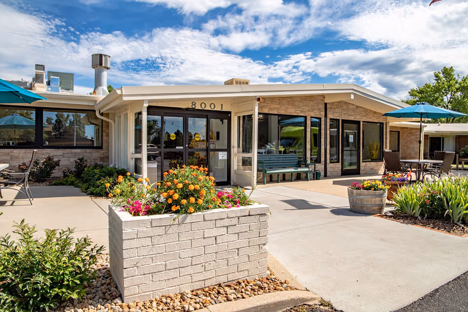 Front entrance of a senior living facility with flower planters, outdoor seating, and '8001' above glass doors.