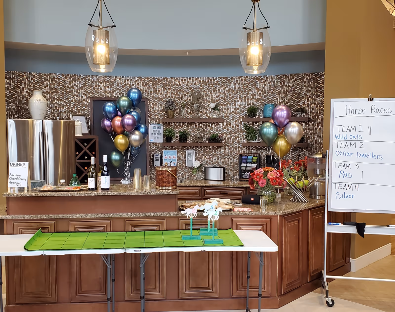 Communal kitchenette and serving counter decorated with balloons, drinks, flowers and a 'Horse Races' whiteboard.