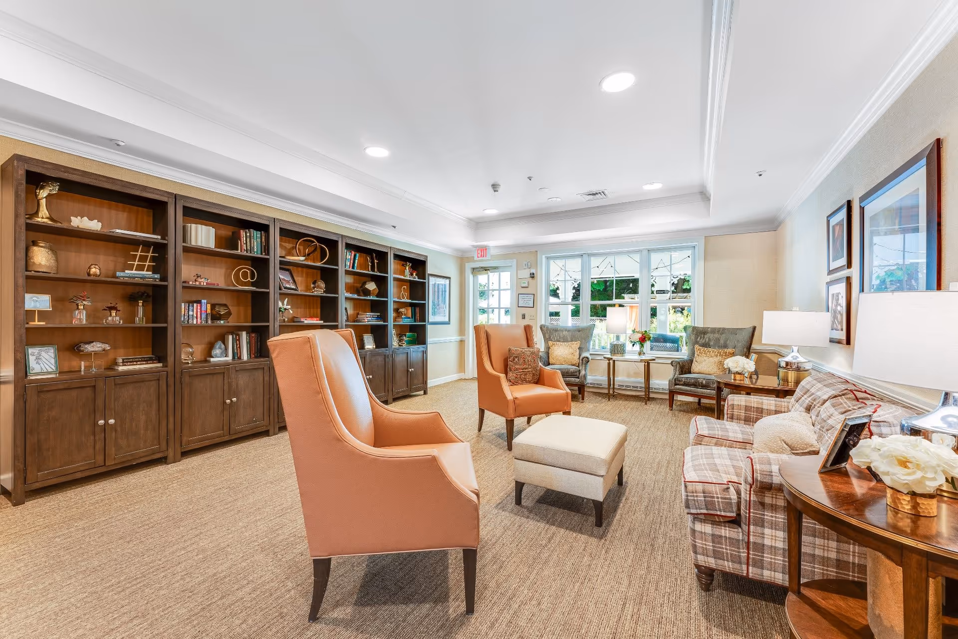 Spacious, bright sitting room with armchairs, a plaid sofa, ottoman, lamps, and built-in wooden bookshelves.