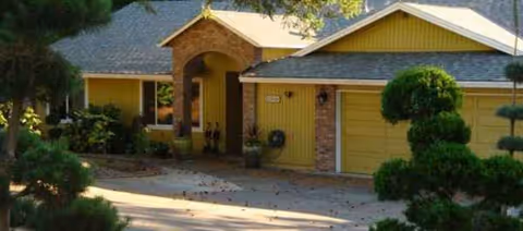 Yellow single-story senior living facility exterior with arched entrance and trimmed shrubs.