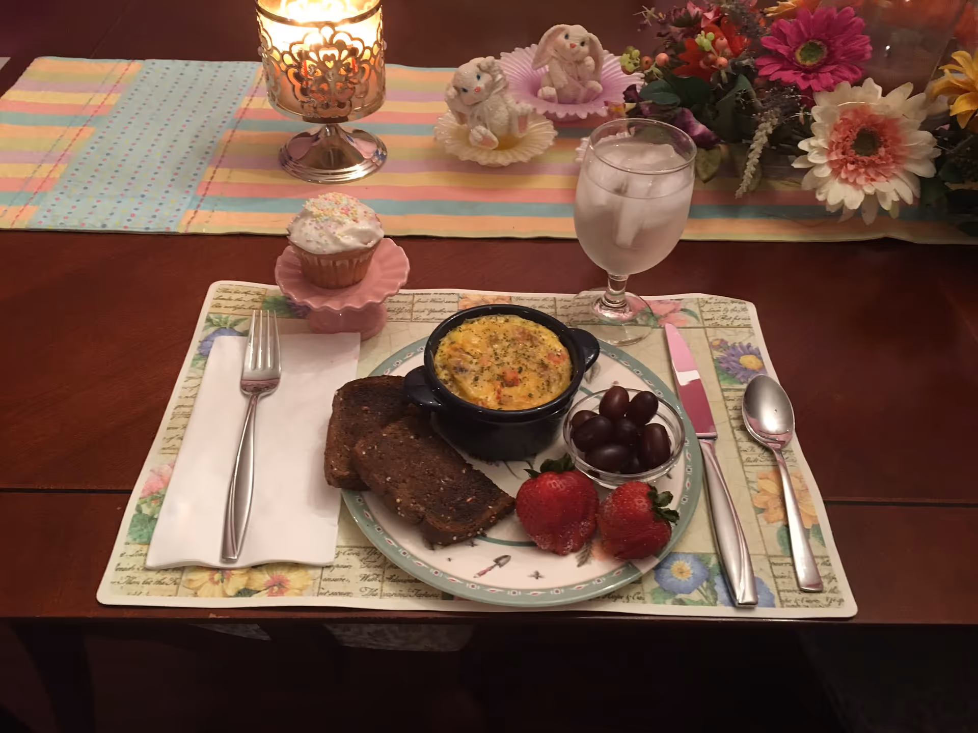 A dining table setting with a plate containing two slices of toasted bread, a small bowl of baked egg dish, a small bowl of grapes, and two strawberries. There is a glass of water with ice, a fork on a napkin, a knife, and a spoon arranged on a floral placemat. A cupcake with white frosting and sprinkles is placed on a pink cupcake stand. In the background, there is a lit candle in a decorative holder and a floral arrangement with colorful flowers and two small bunny figurines.
