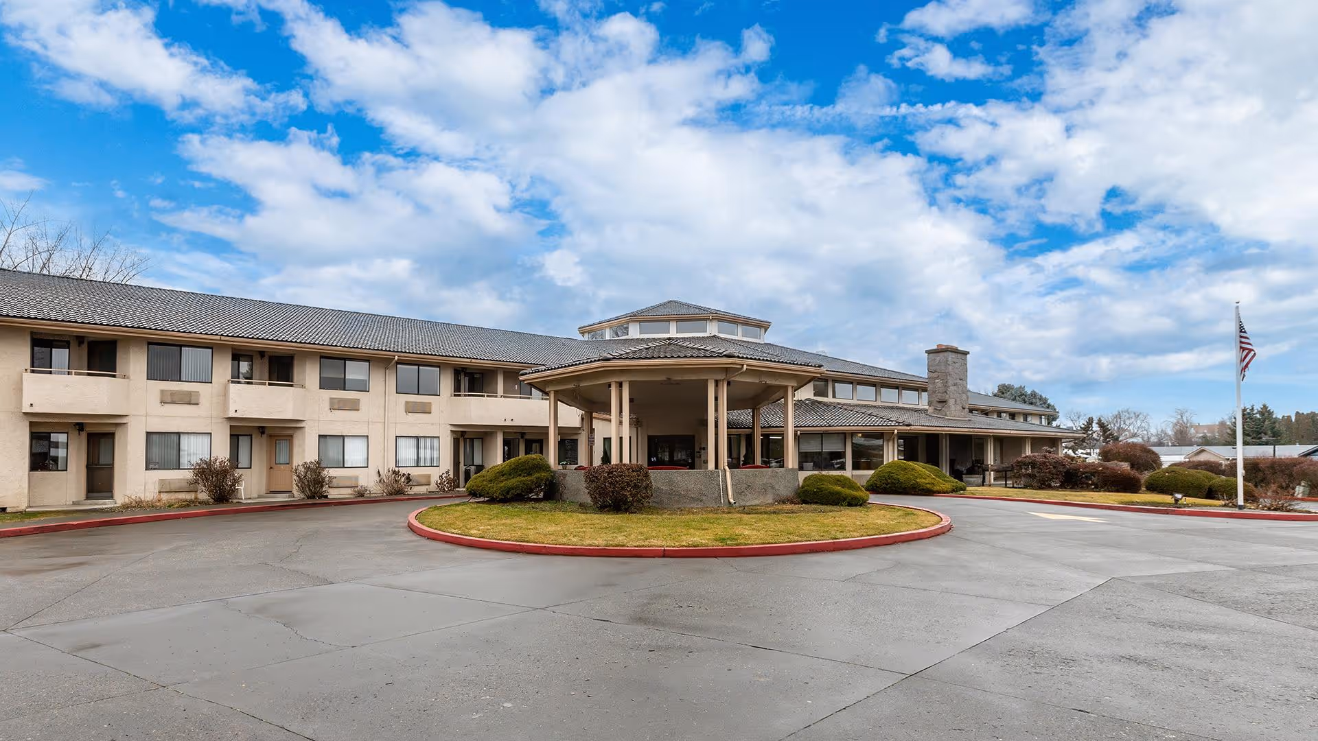 Exterior view of a two-story senior living facility building with a covered entrance, landscaped bushes, and an American flag on a flagpole under a partly cloudy sky.