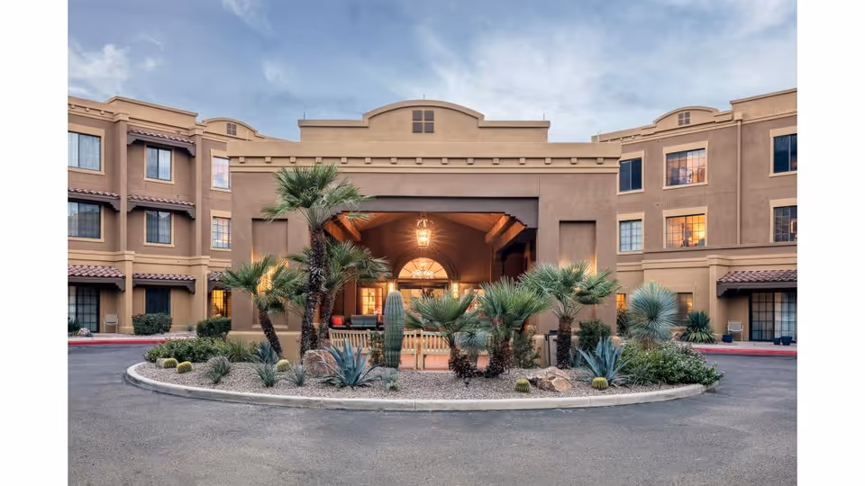 Front exterior view of a senior living facility with a circular driveway and desert landscaping featuring palm trees, cacti, and other desert plants. The building has a tan stucco finish with multiple windows and a covered entrance with warm lighting inside.