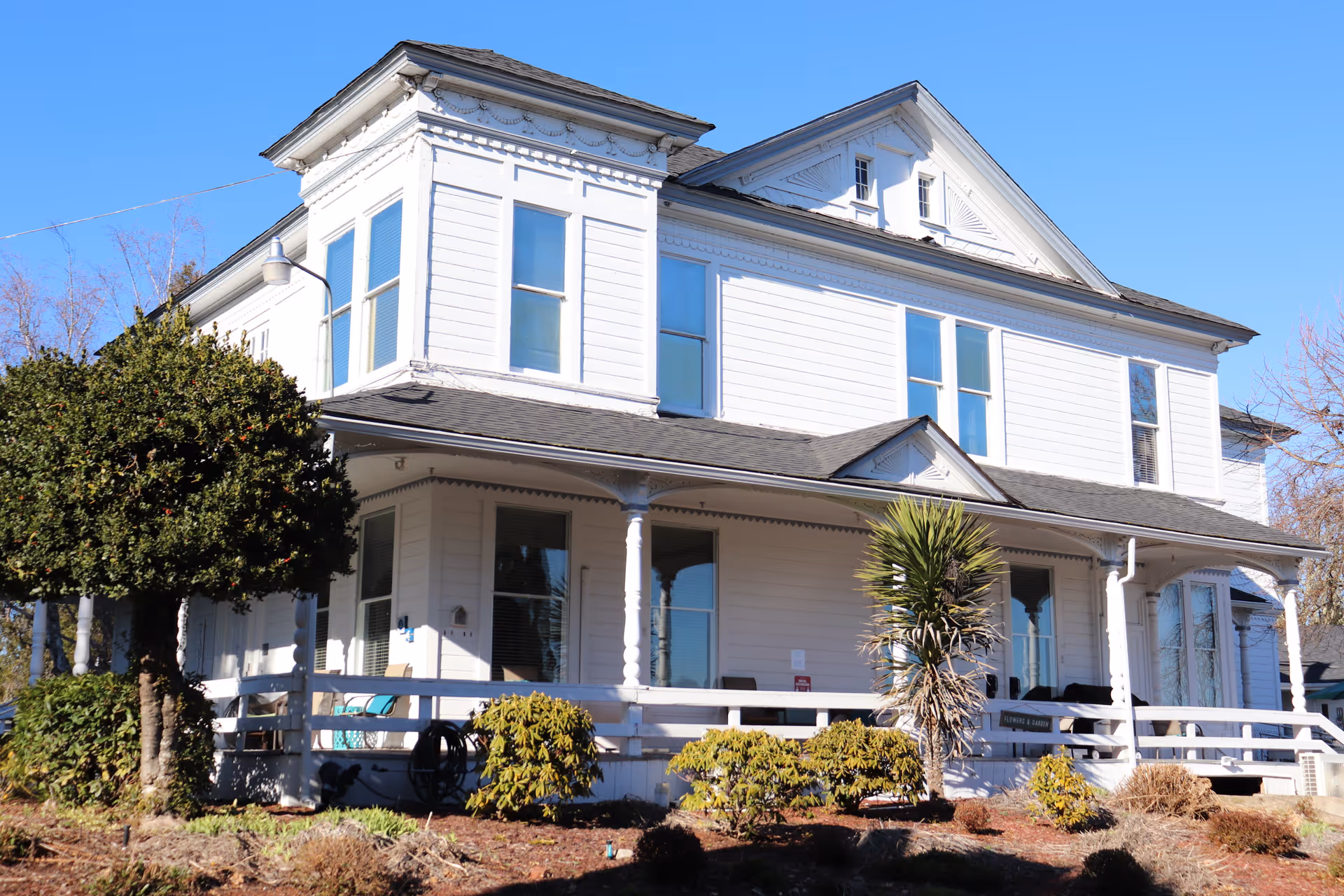 Exterior view of a large, white two-story house with multiple windows and a wrap-around porch, surrounded by shrubs and small trees under a clear blue sky.