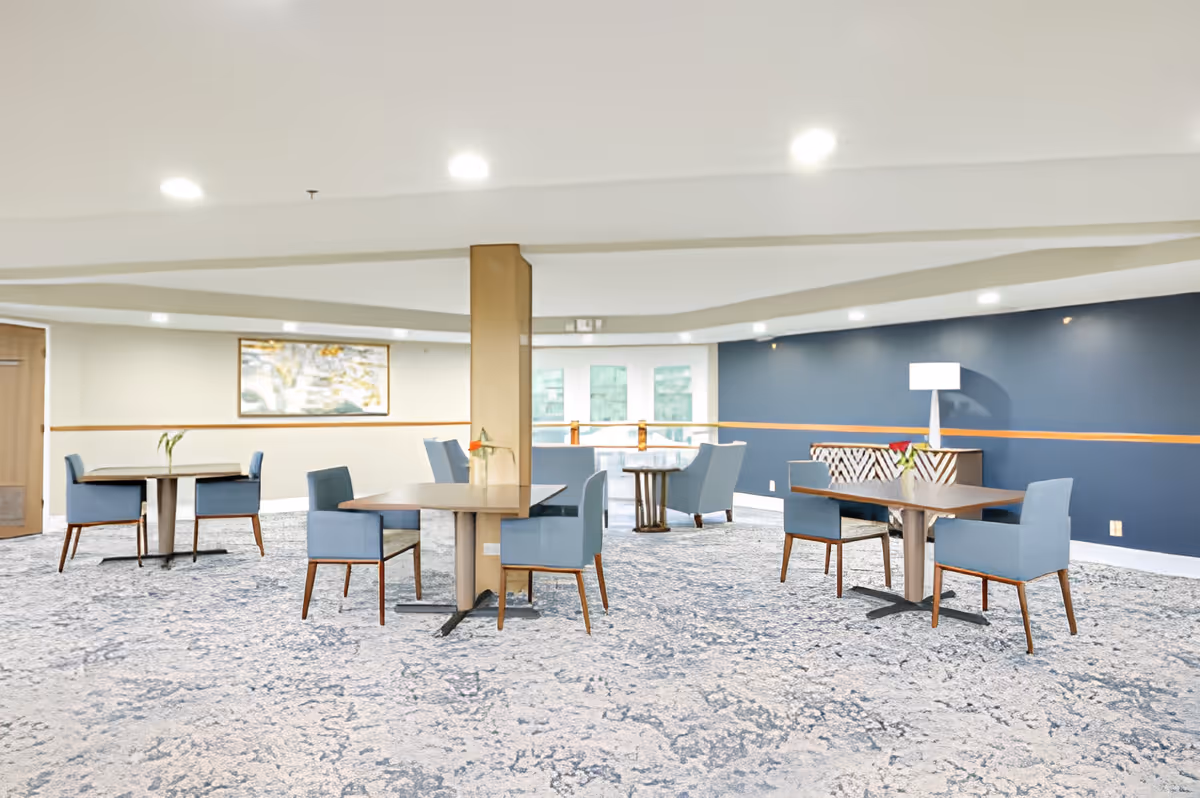 A spacious, well-lit common area with several tables and blue upholstered chairs arranged for seating. The room features a patterned carpet, a blue accent wall with a lamp and a sideboard, and a large window letting in natural light.