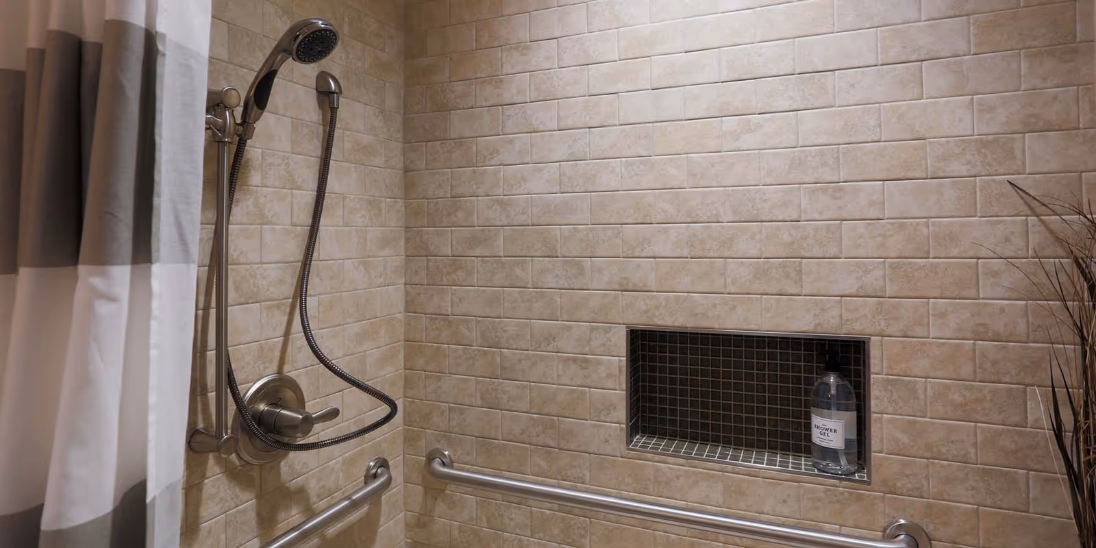 A tiled shower area with beige rectangular tiles, a handheld showerhead attached to a vertical bar, two horizontal grab bars, a built-in recessed shelf holding a bottle of shower gel, and a gray and white shower curtain partially visible on the left side.