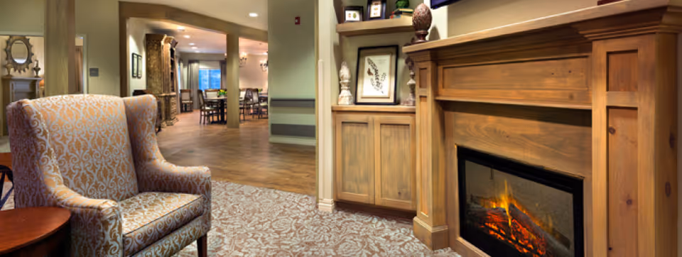 Cozy interior of a senior living facility featuring a patterned armchair next to a wooden electric fireplace with a mantle displaying decorative items and framed artwork. The room has carpeted flooring near the fireplace and wooden flooring extending into a dining area with tables and chairs in the background.
