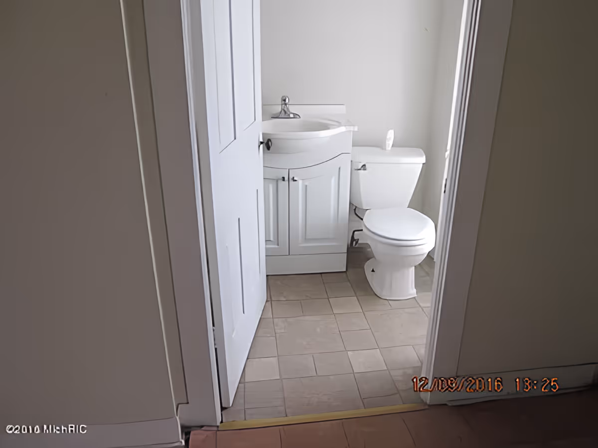 View into a small bathroom with a white door partially open, showing a white sink with cabinet and a white toilet. The floor is tiled and the walls are painted white.