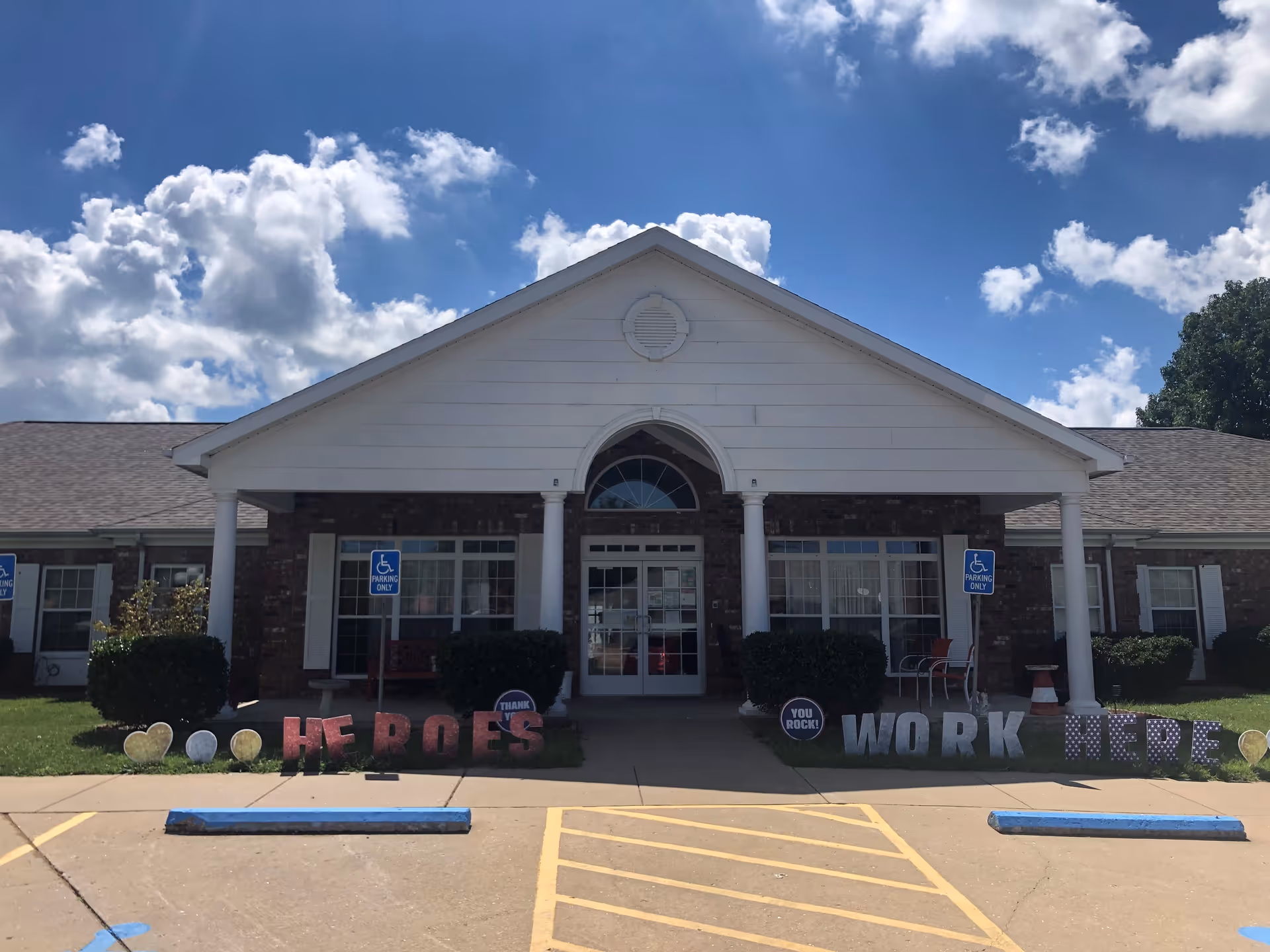 Front exterior view of a single-story brick building with white columns and a triangular white pediment above the entrance. There are two handicap parking signs and spaces in front of the building. Decorative letters on the lawn spell out 'HEROES WORK HERE' with additional signs saying 'THANK YOU' and 'YOU ROCK!'. The sky is blue with scattered clouds.