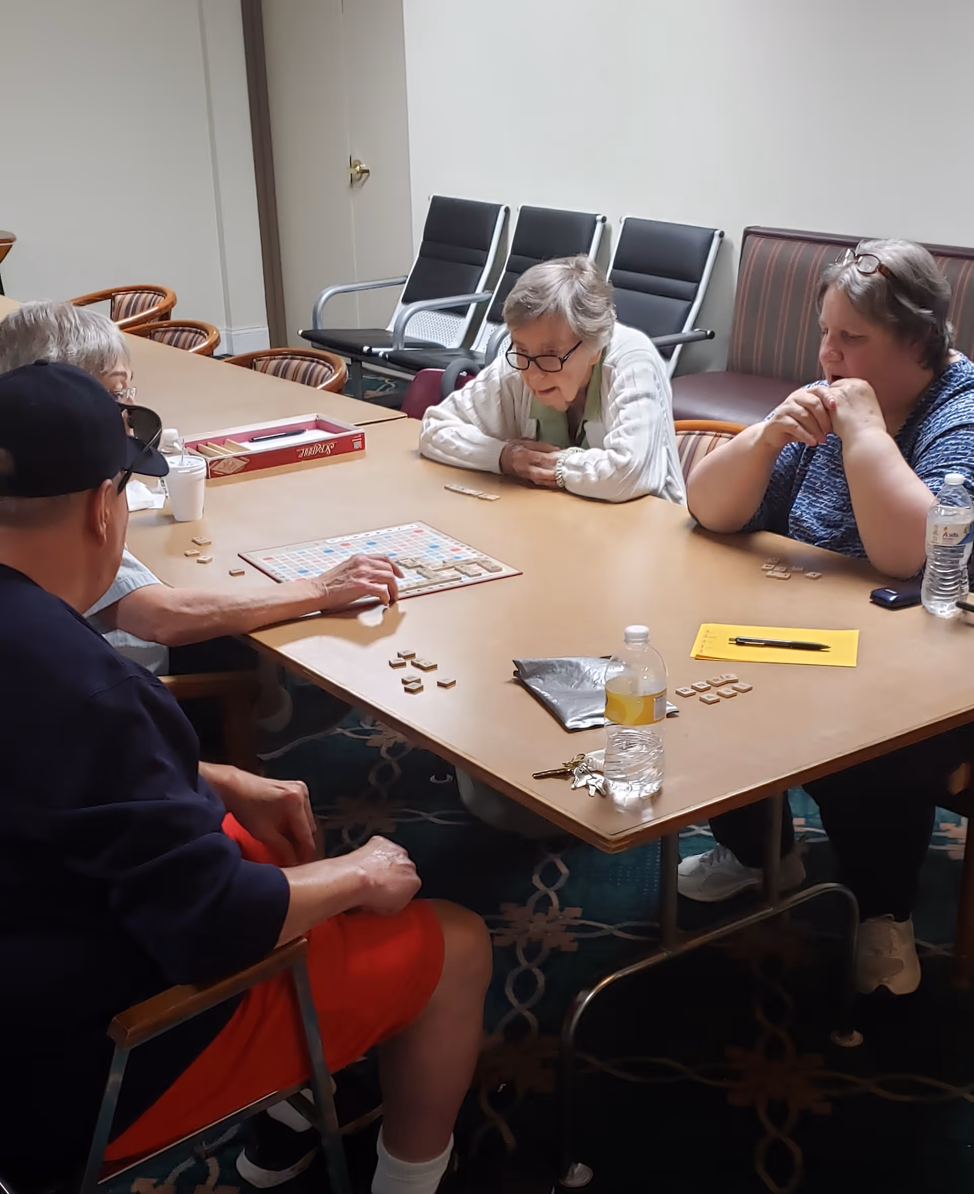 Four elderly people sitting around a rectangular table playing a board game resembling Scrabble in a room with chairs and a patterned carpet.
