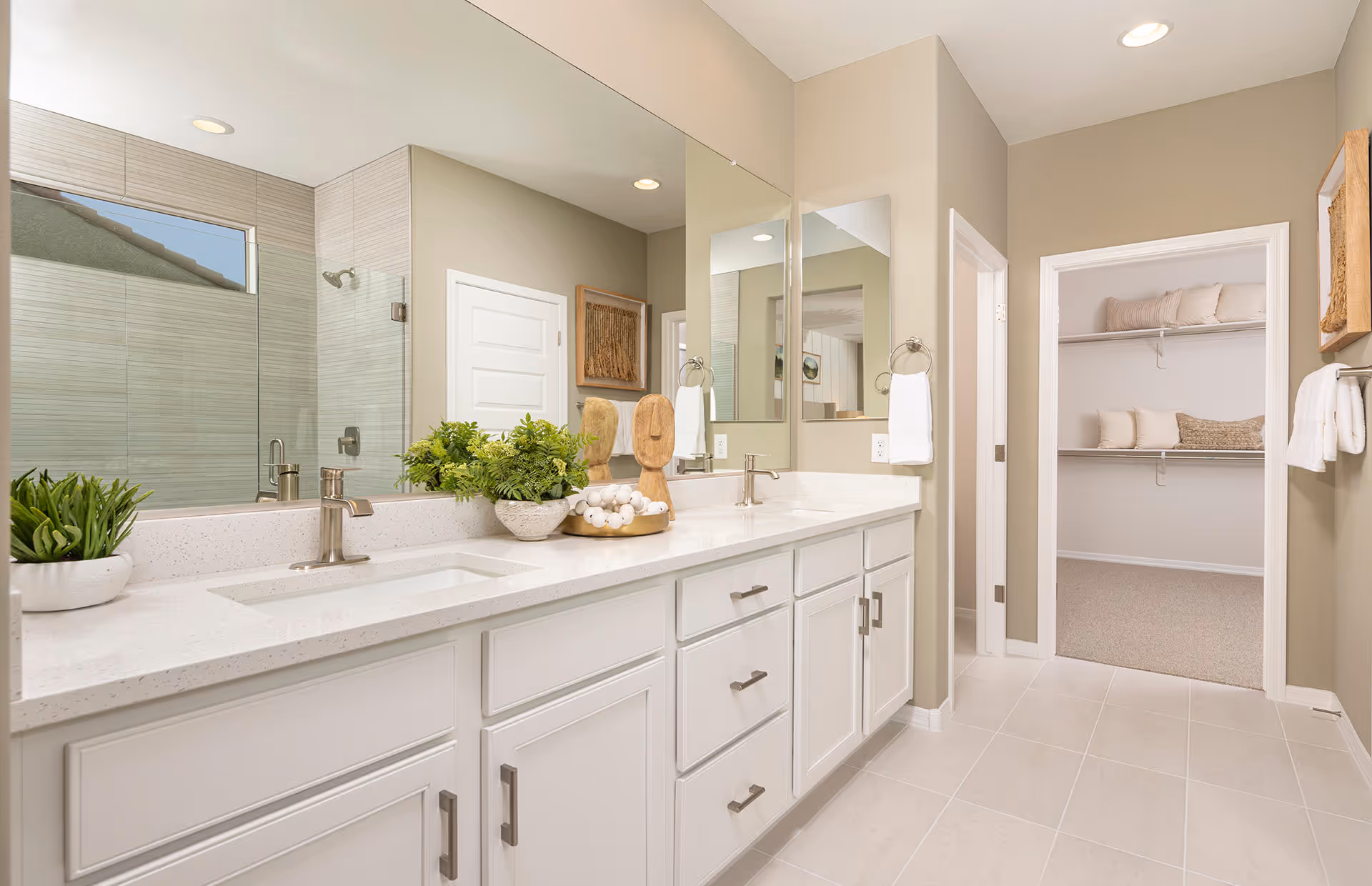 Bright and modern bathroom with a long white vanity featuring two sinks and silver faucets. A large mirror spans the length of the vanity. There are decorative plants and wooden sculptures on the countertop. To the right, an open doorway leads to a walk-in closet with shelves holding pillows and blankets. The walls are painted beige, and the floor is tiled in light colors.