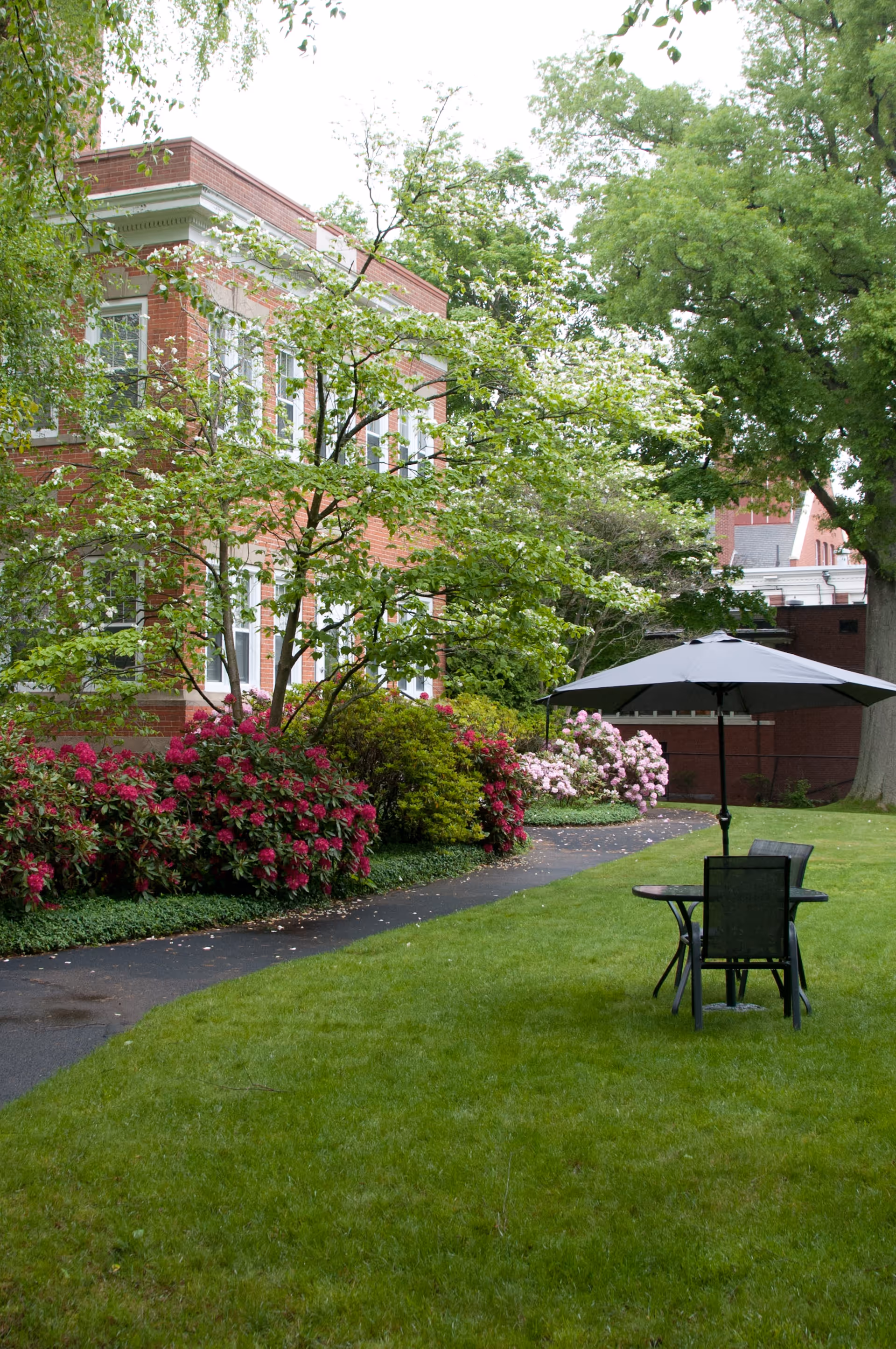 Brick building beside a green lawn and flowering garden with a pathway and a patio table with an umbrella.