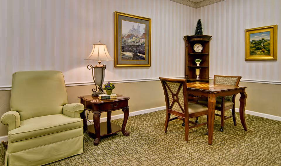 A cozy sitting area in a senior living facility featuring a green upholstered armchair, a wooden side table with a lamp and small plant, a wooden table with two chairs, a corner shelf with decorative items, and two framed paintings on the walls with striped wallpaper.