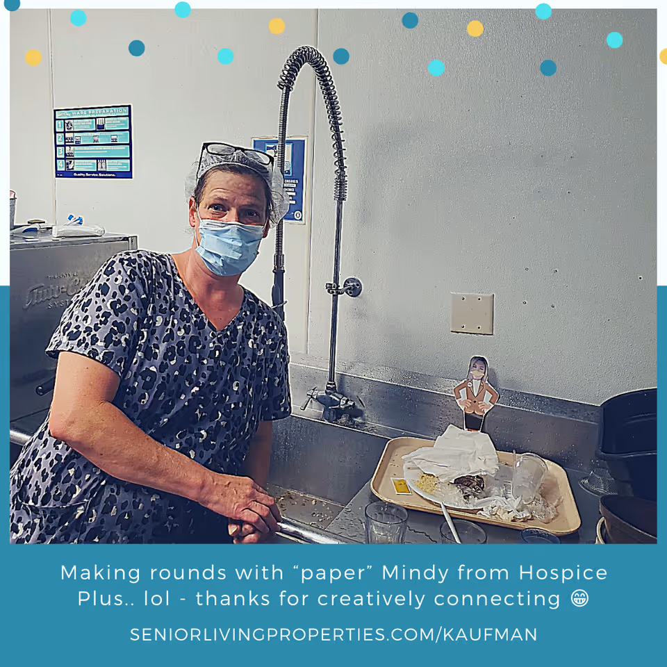 A masked staff member in scrubs standing at a stainless steel sink and dishwashing area with a small paper doll on a tray.