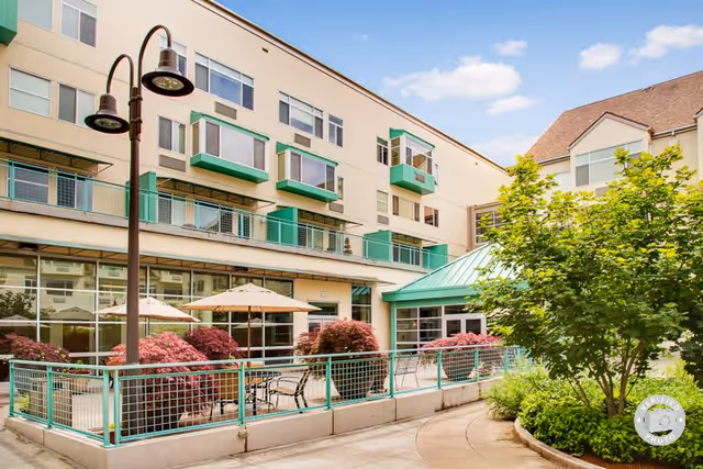 Outdoor patio area of a senior living facility with tables, chairs, umbrellas, and landscaped greenery including bushes and a tree, adjacent to a multi-story building with balconies and large windows under a partly cloudy sky.