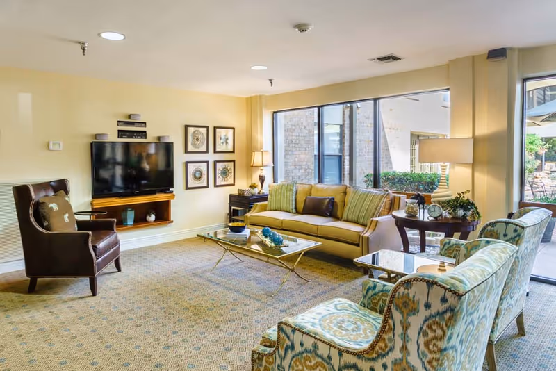 A bright and cozy living room with a beige sofa, two patterned armchairs, a glass coffee table, a side table with a lamp, and a wall-mounted flat-screen TV. Large windows provide a view of an outdoor patio area with greenery.