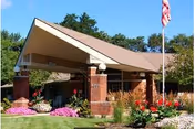 Front entrance of a brick senior living building with a pitched canopy, landscaped flower beds and an American flag.