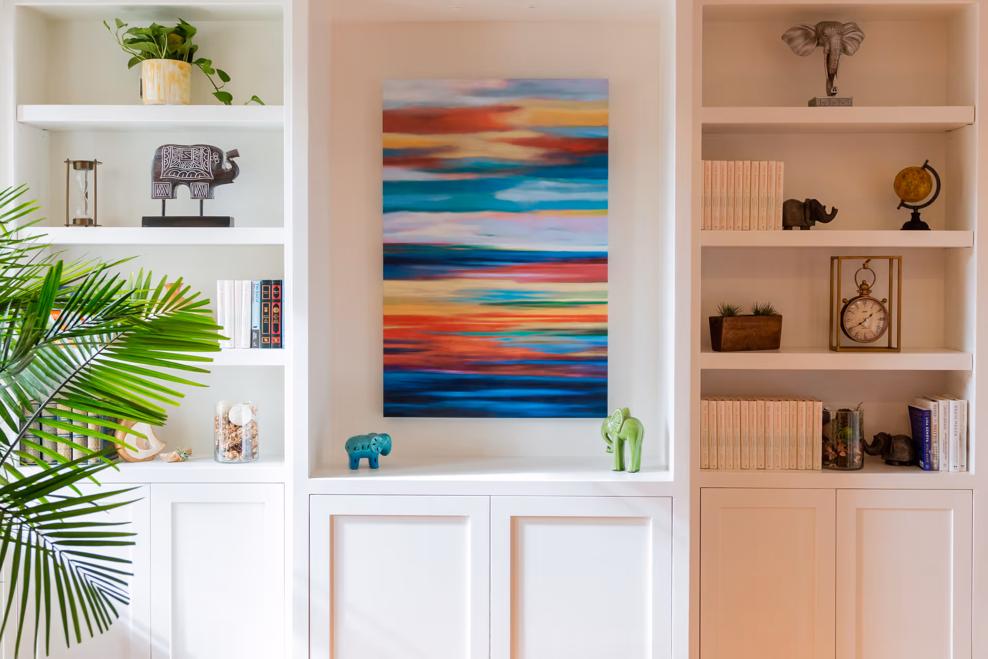 A white built-in shelving unit with various decorative items including small elephant figurines, books, a clock, a globe, and plants. In the center is a colorful abstract painting with horizontal streaks of red, blue, orange, and green. A green leafy plant is partially visible on the left side.