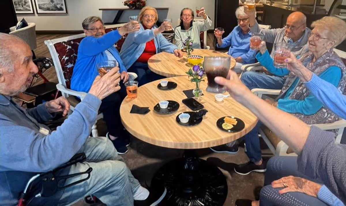 A group of elderly people sitting around two round wooden tables in a common area, raising their glasses in a toast. The tables have small plates with snacks, napkins, and small flower vases. The setting appears warm and social.