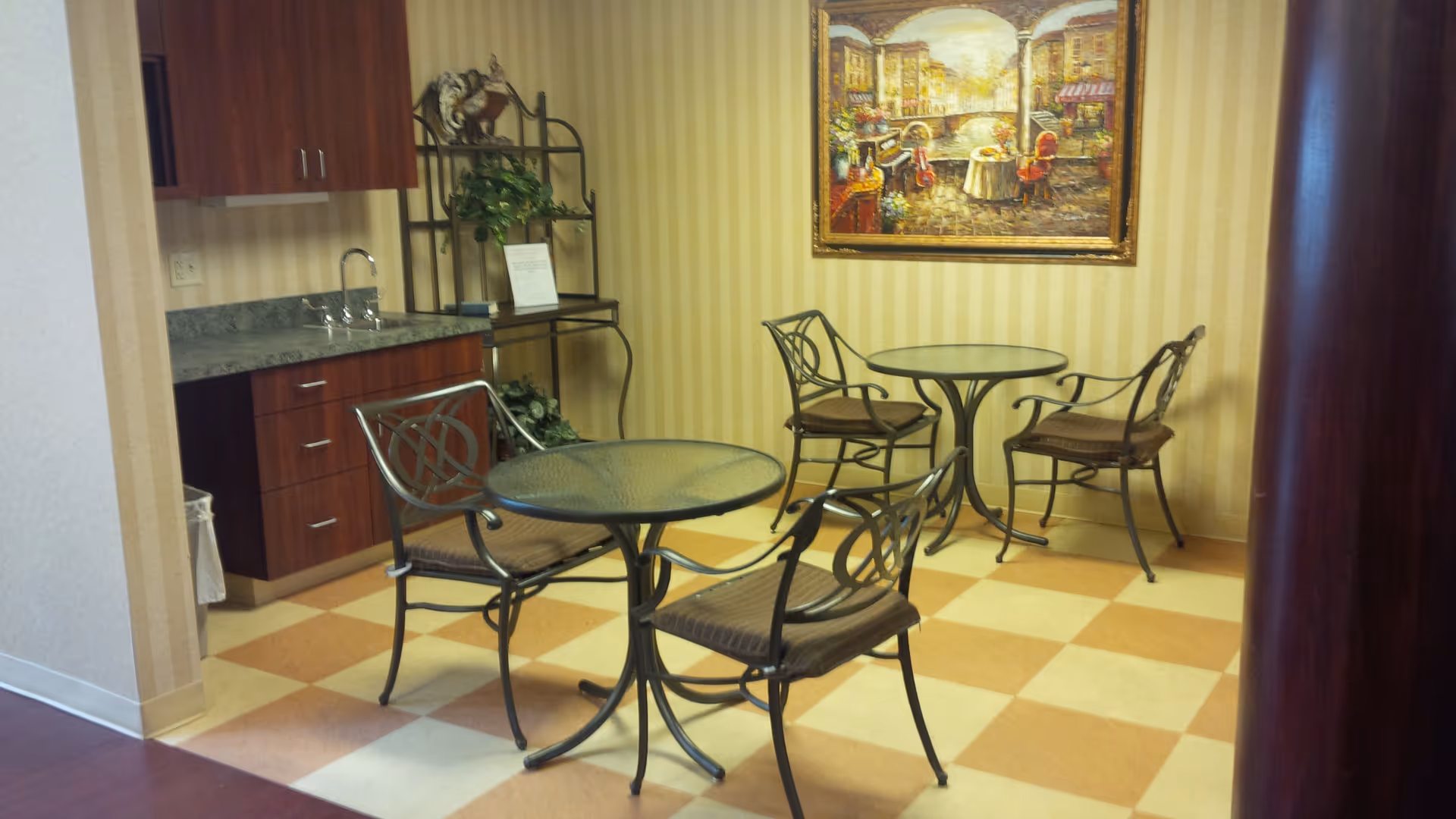 Small seating area with two round glass-top tables and four metal chairs with cushions, located next to a kitchenette with wooden cabinets and a sink. A decorative metal shelf with plants and a framed painting of an outdoor cafe scene hang on the striped wallpapered wall.